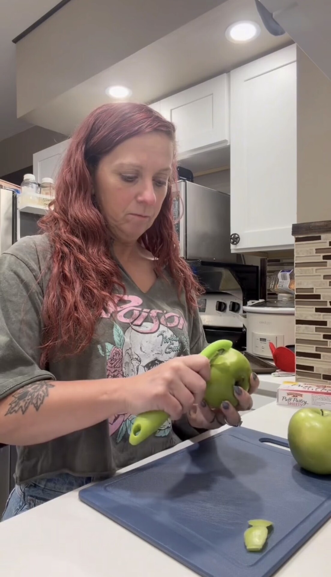 A woman peeling a green apple with a vegetable peeler in a kitchen.