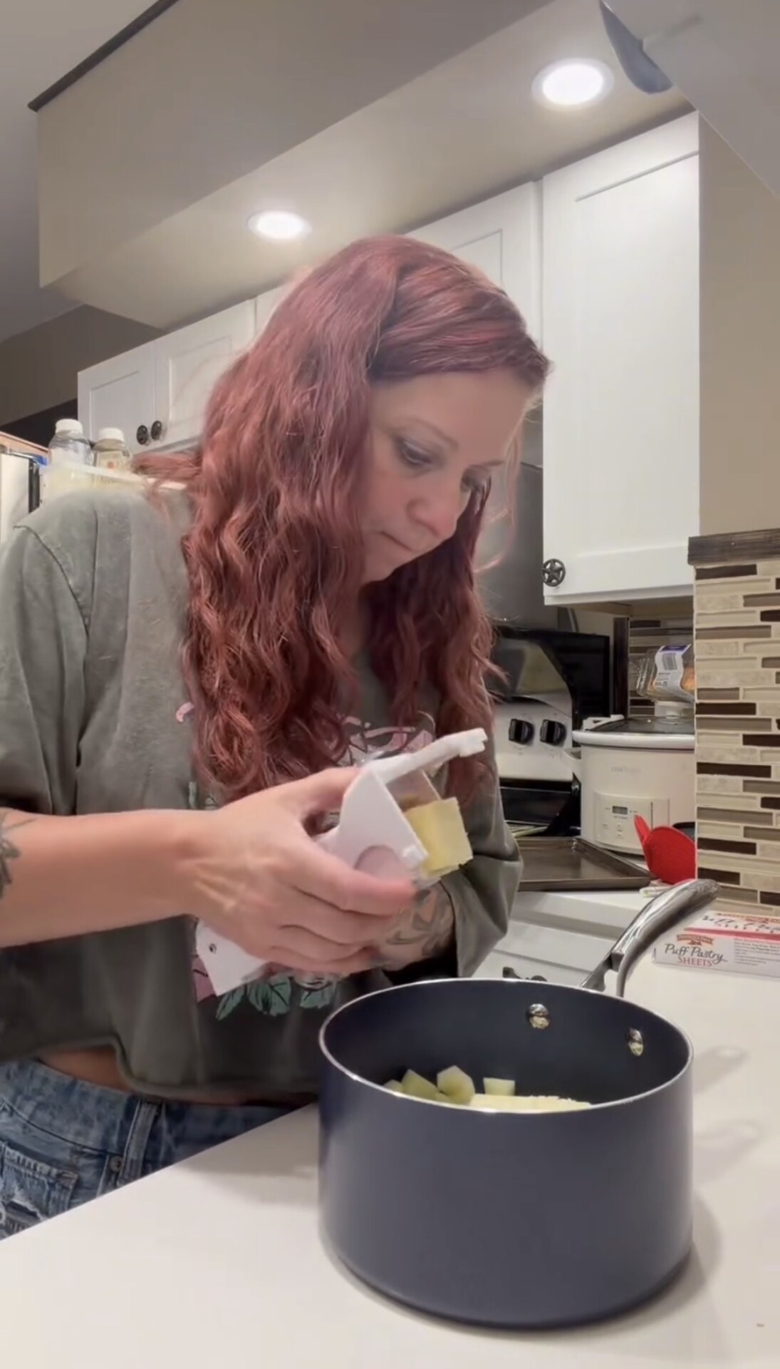 A woman uses a handheld vegetable slicer to cut potatoes into a dark cooking pot on a kitchen counter.