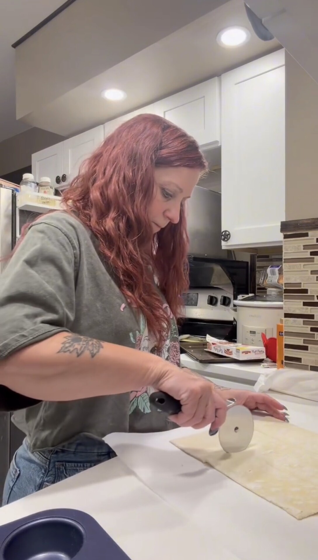 A woman with reddish hair uses a round pizza cutter to slice a sheet of dough on a kitchen counter.