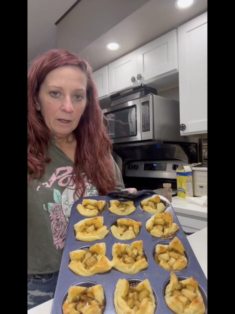 A woman in a kitchen holds a muffin tin filled with freshly baked apple pastries.