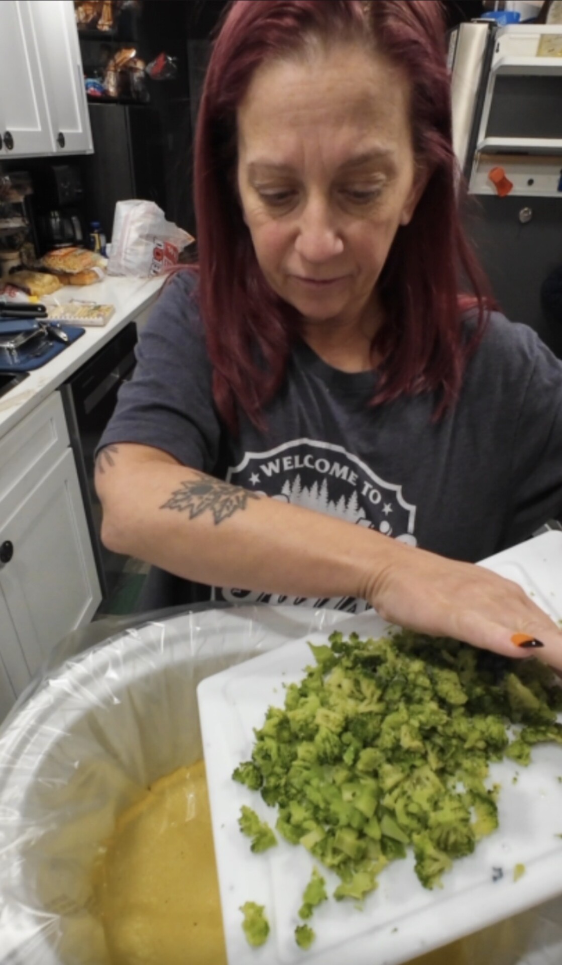 A woman with red hair empties chopped broccoli from a white cutting board into a slow cooker.