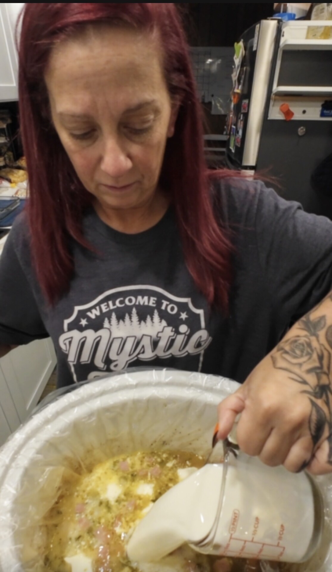 A woman wearing a Welcome to Mystic t-shirt pours a white liquid from a measuring cup into a slow cooker containing a mixture of ingredients.