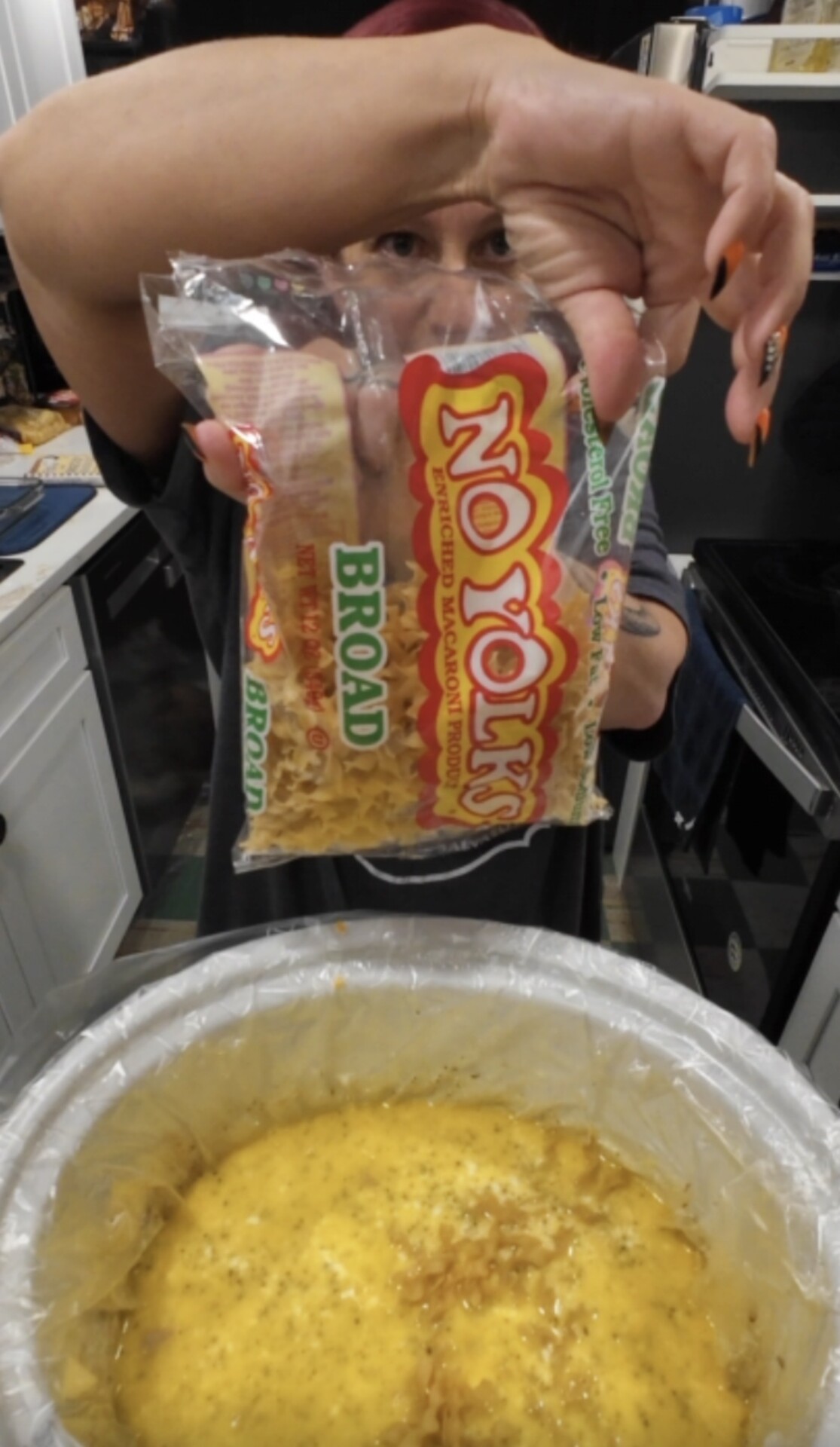 A person holds a bag of No Yolks broad enriched macaroni product over a slow cooker filled with a creamy, cheesy dish.
