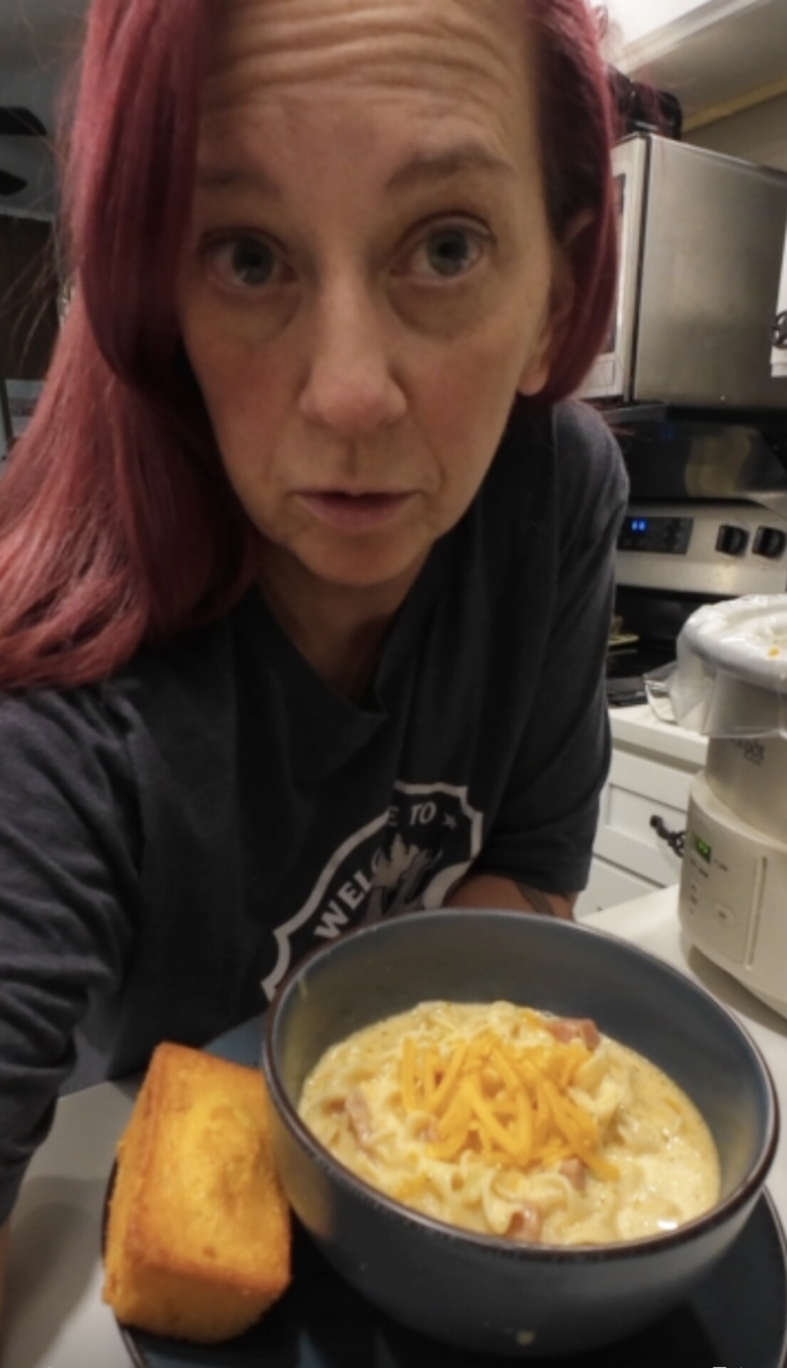 A person with red hair stands behind a bowl of creamy soup topped with shredded cheddar cheese and a piece of cornbread.