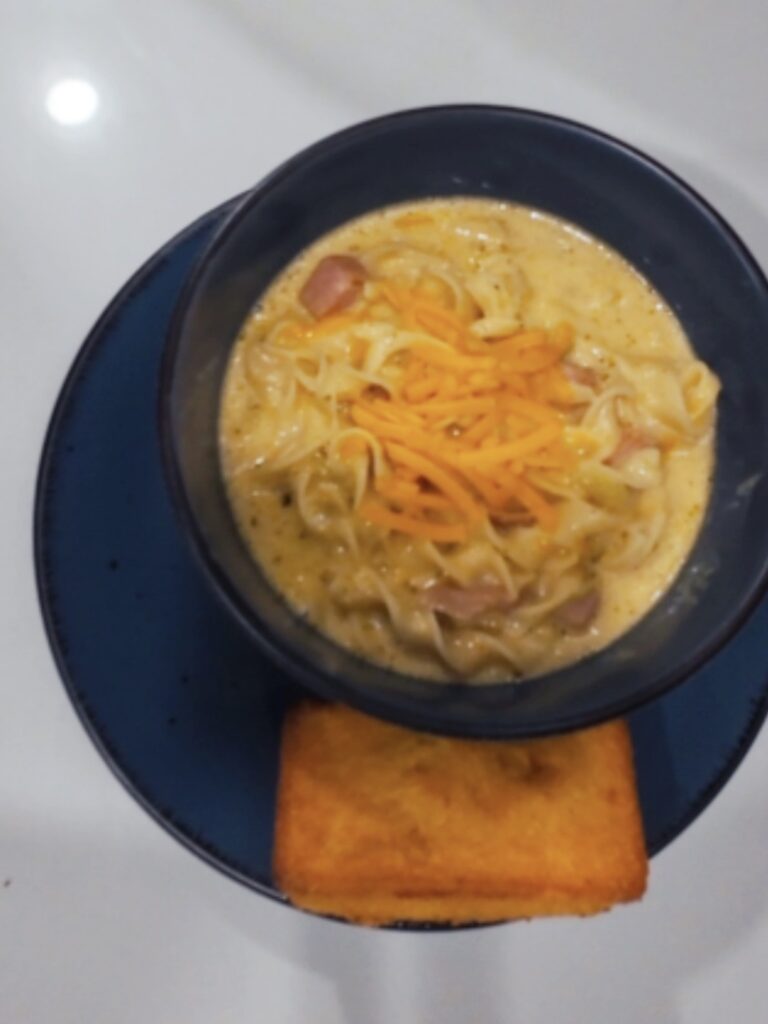 A bowl of creamy noodle soup topped with shredded cheddar cheese, served with a piece of cornbread.