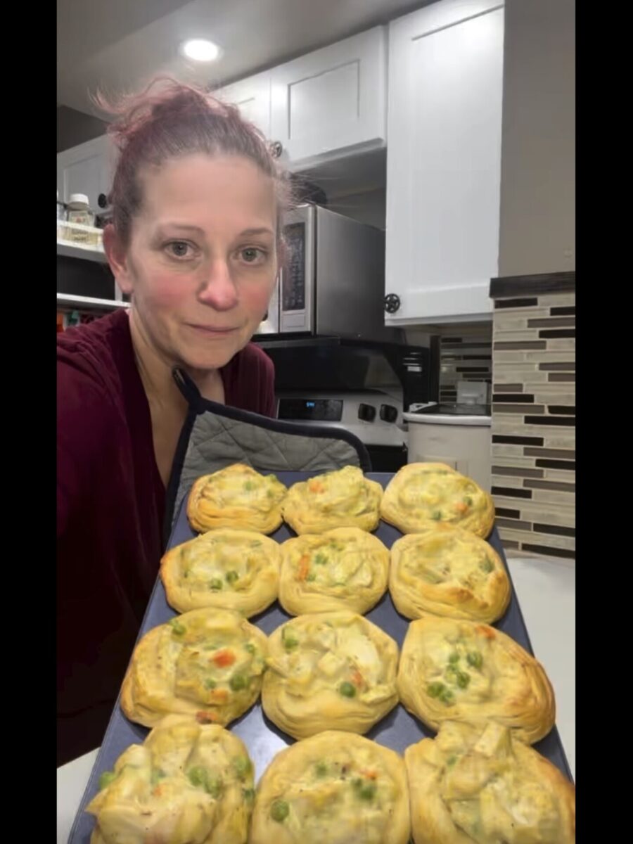 A woman holds a baking sheet of twelve savory chicken and vegetable pastries.