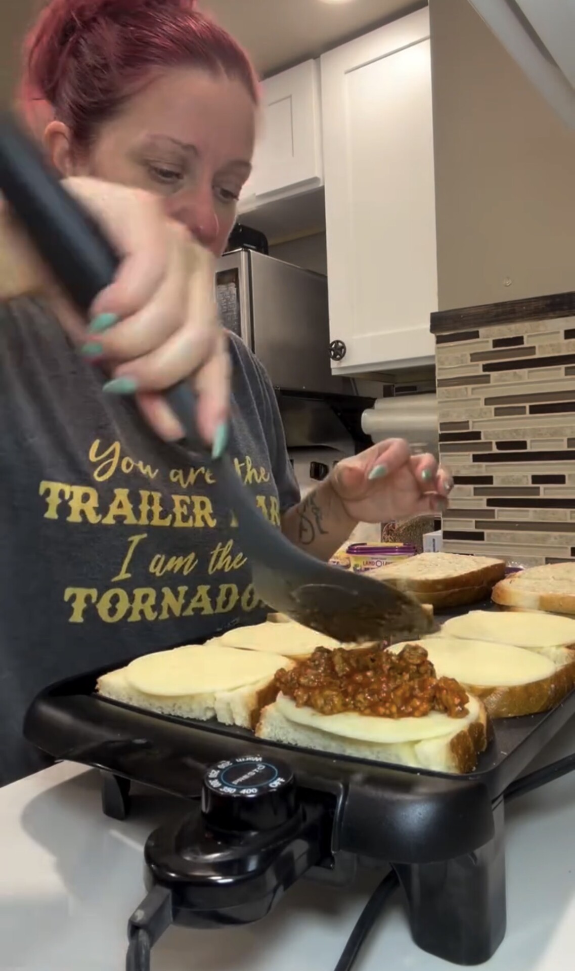 A person uses a spatula to spread sloppy joe meat onto slices of bread topped with cheese on an electric griddle.
