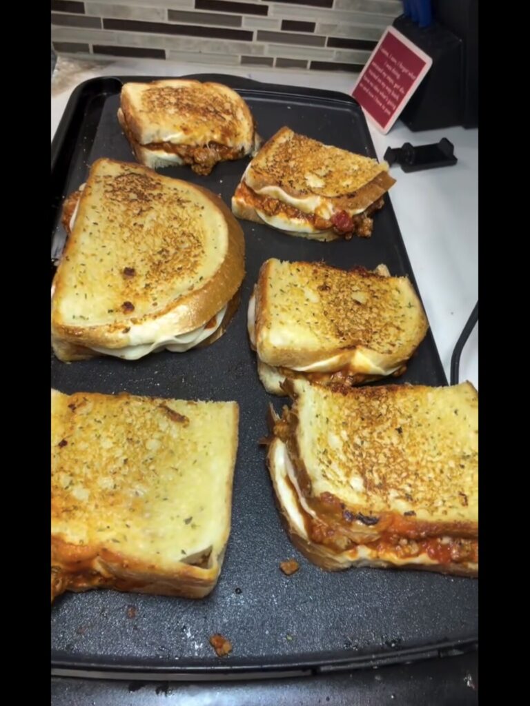 Five grilled sandwiches with melted cheese and red sauce cooking on a flat electric griddle.
