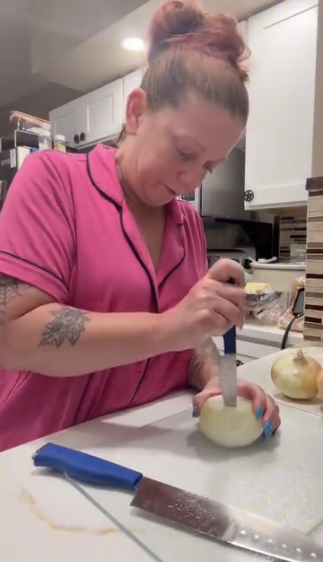 A woman in pink pajamas uses a knife to core an onion on a cutting board.