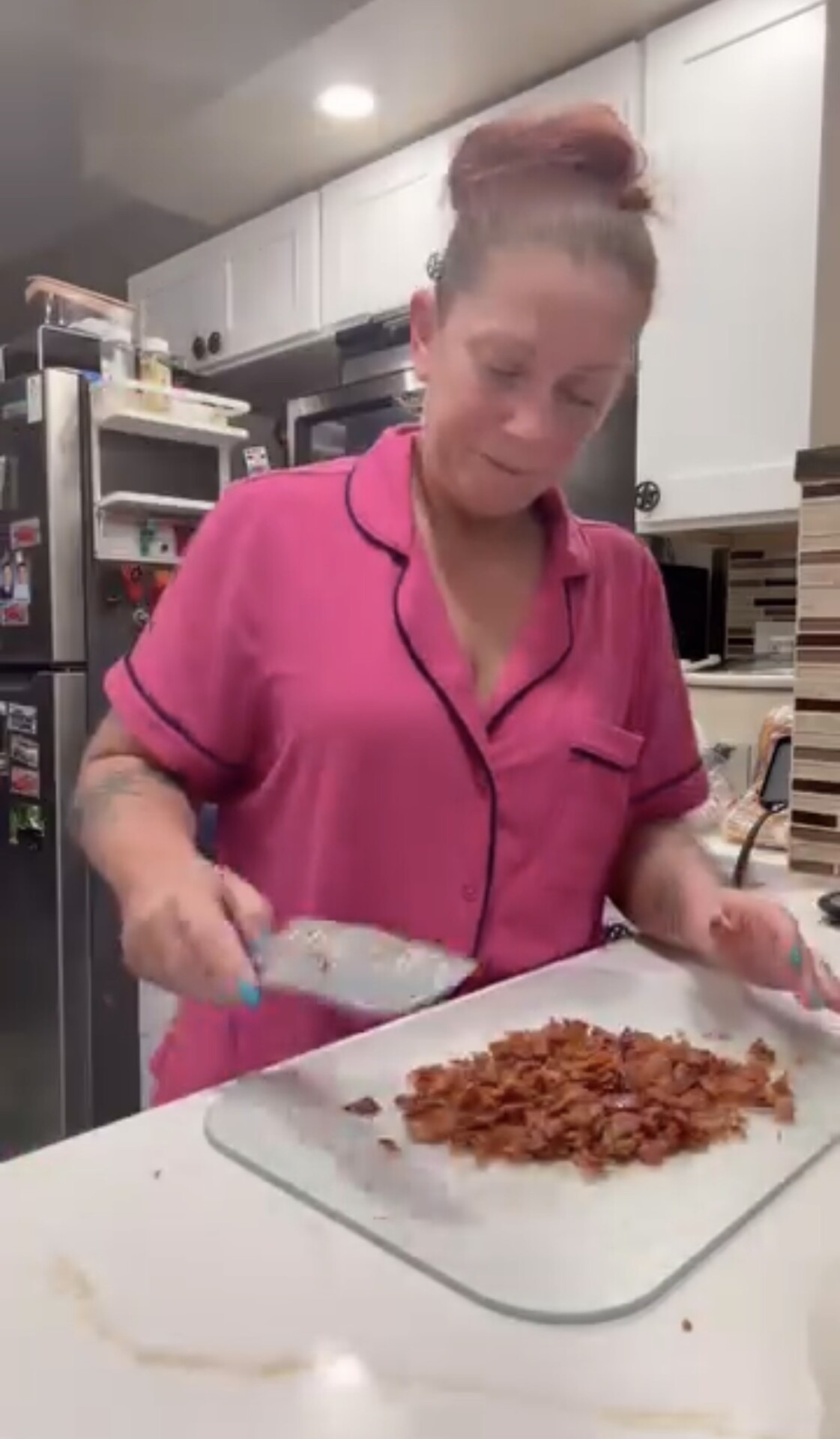 A woman in pink pajamas chops cooked bacon on a cutting board in a kitchen.