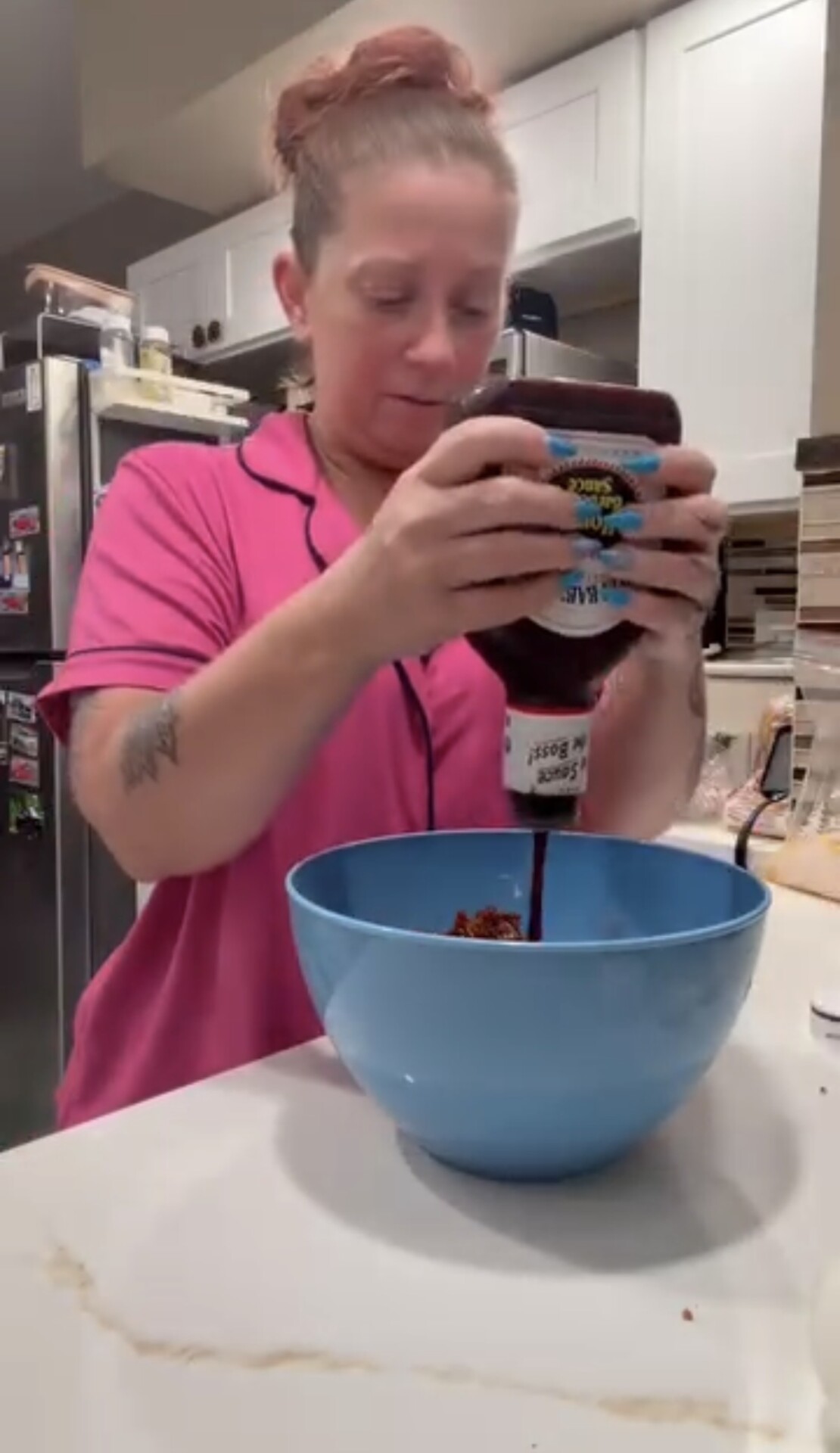 A woman in a pink shirt squeezes a bottle of barbecue sauce into a blue mixing bowl.