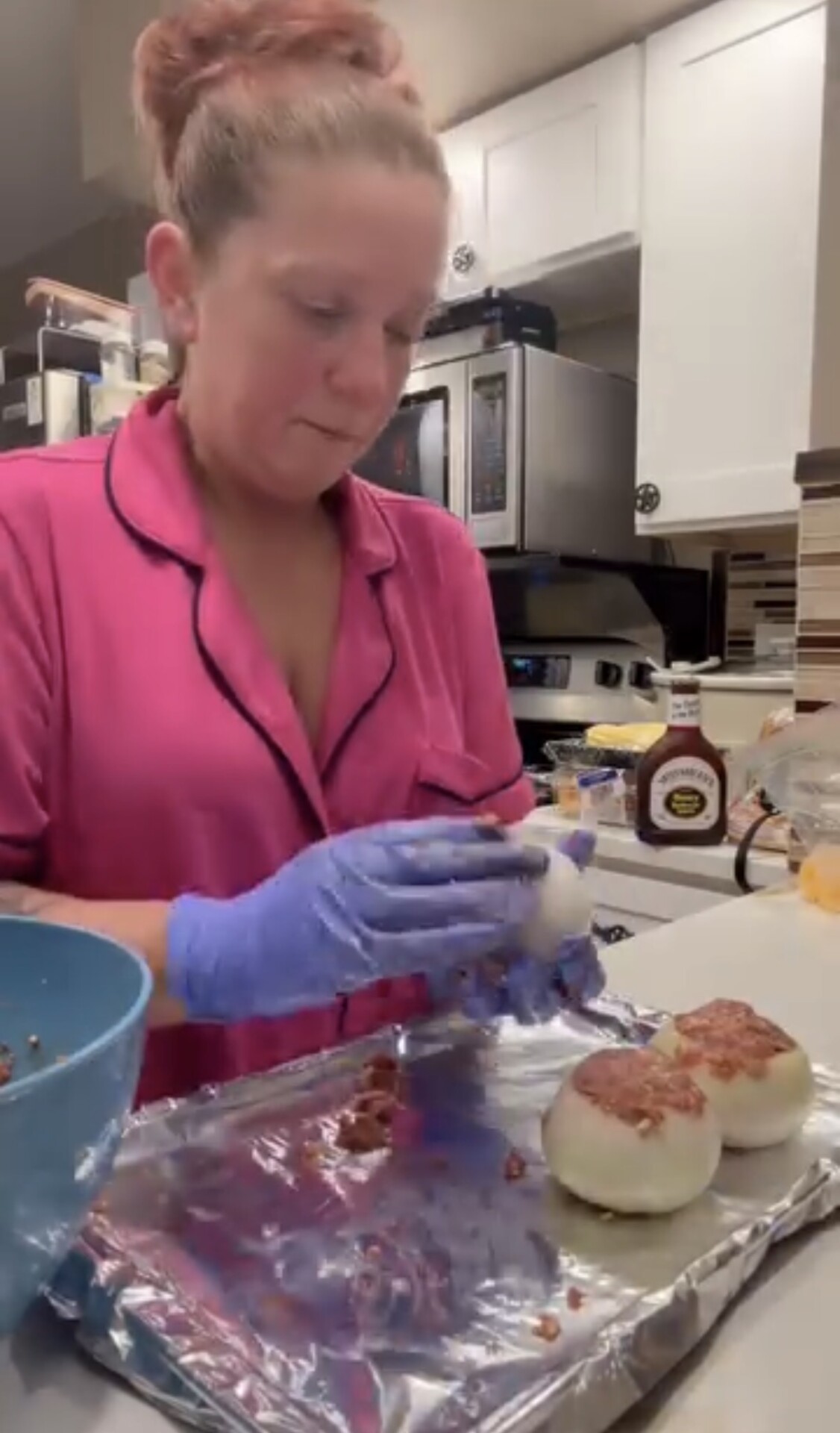 A woman wearing a pink pajama top and blue gloves stuffs meat into hollowed-out onion halves on a foil-lined baking sheet.