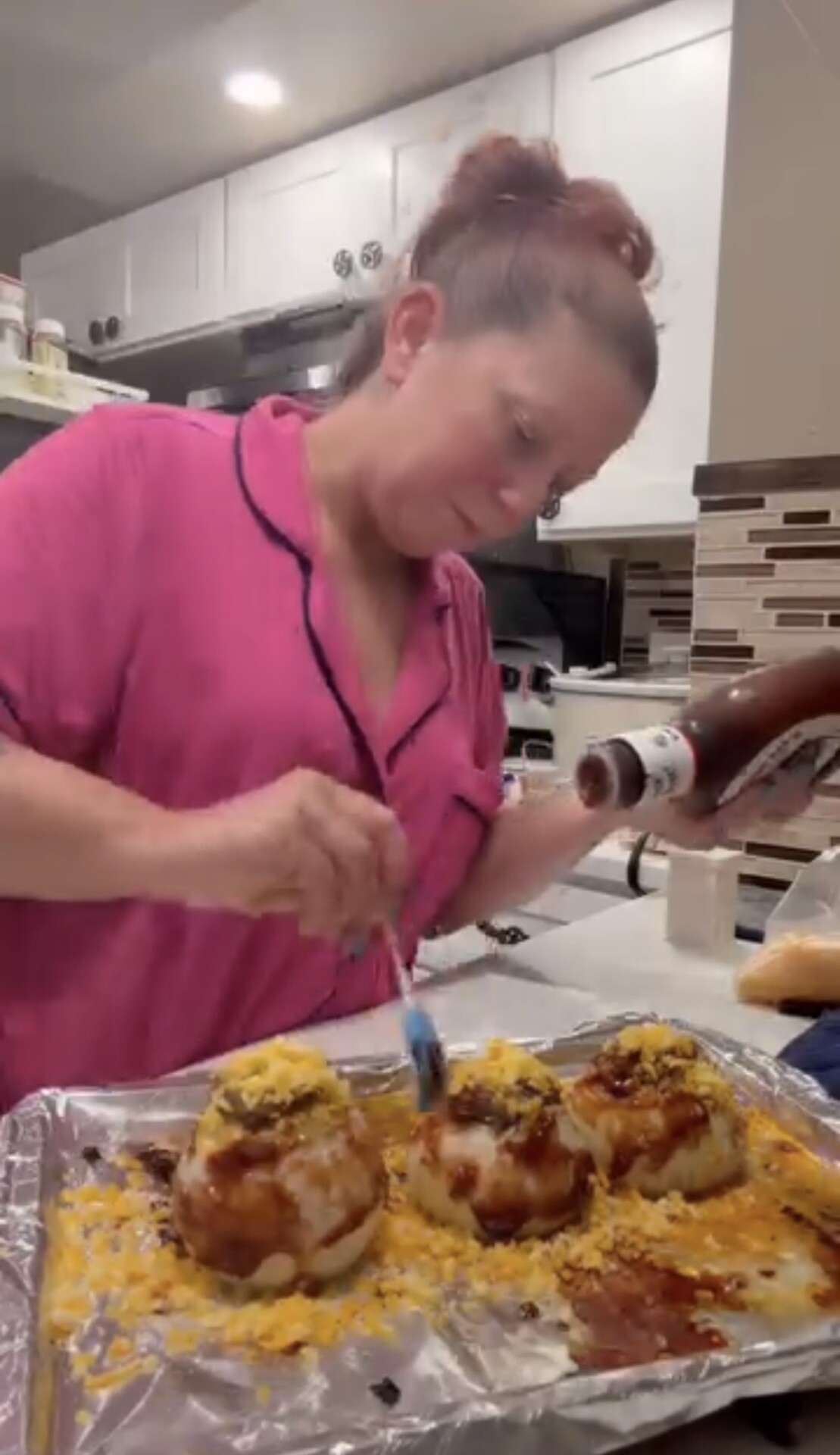 A woman in pink pajamas brushes barbecue sauce onto three whole onions topped with shredded cheese on a foil-lined baking sheet.