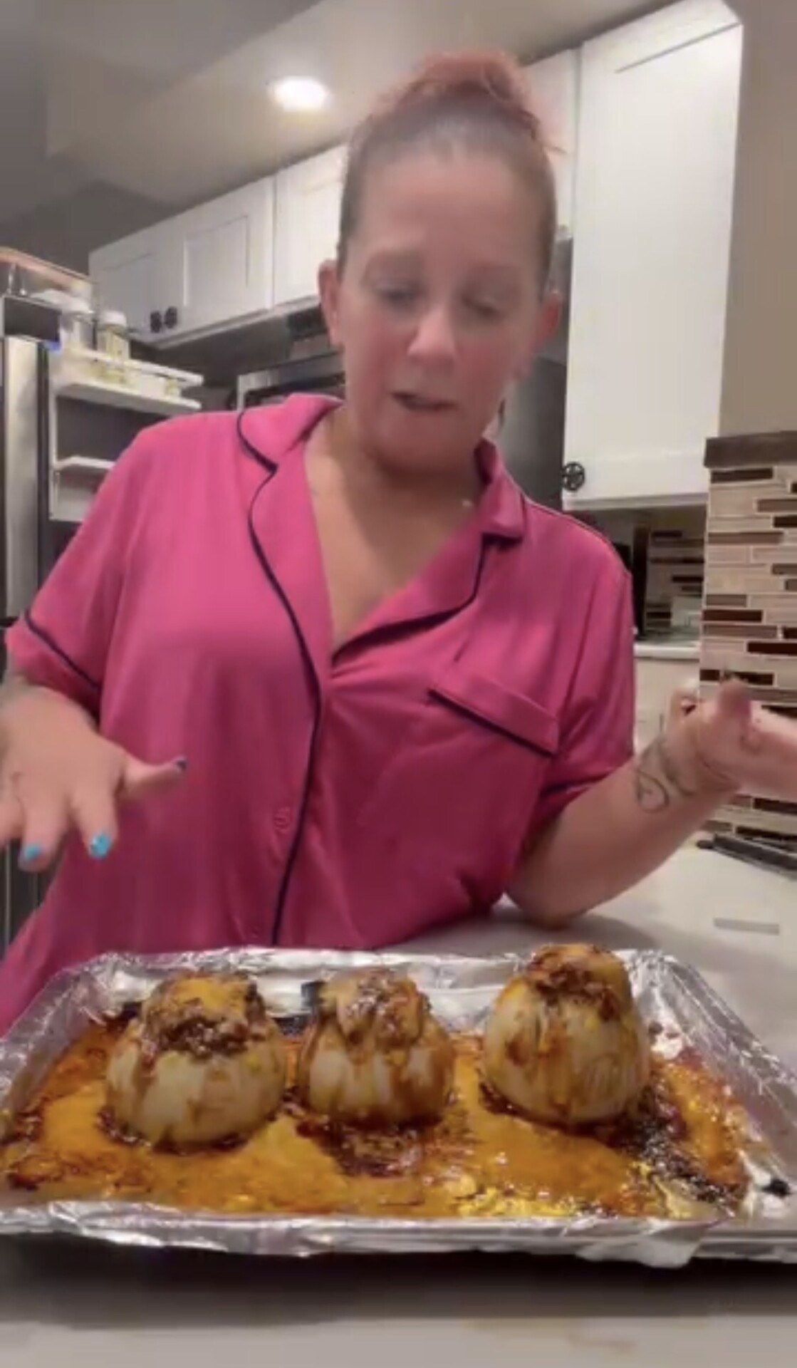 A person in pink pajamas stands behind a baking sheet holding three baked, stuffed onions topped with melted cheese.