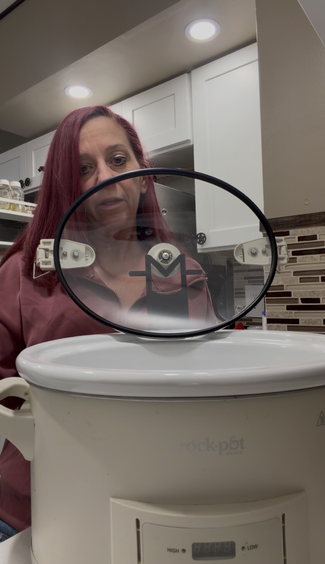 A woman holding a circular glass Crock-Pot lid over the open base of the appliance.