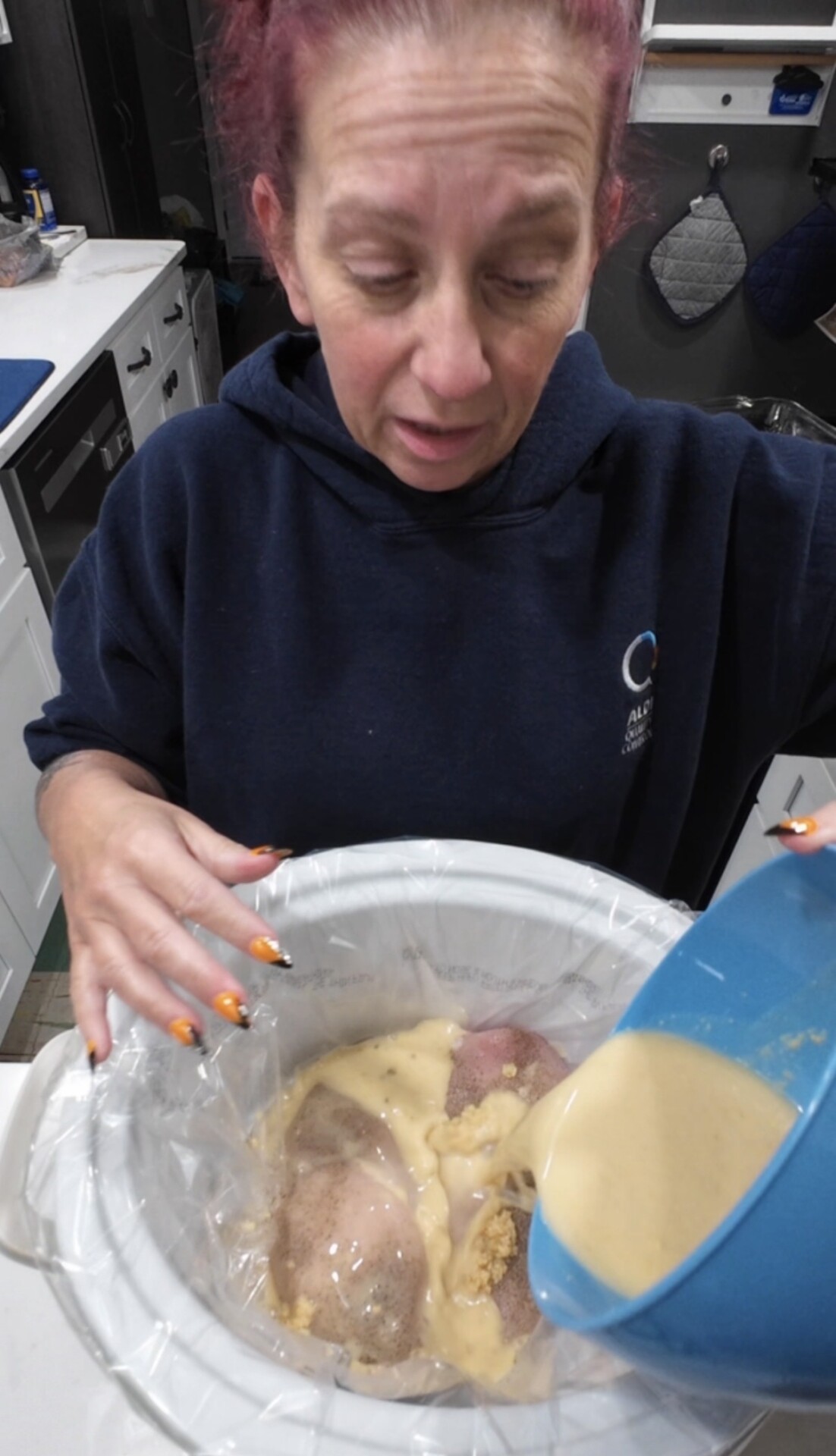 A person in a blue hoodie pours a yellow liquid from a blue bowl into a slow cooker containing raw chicken breasts.