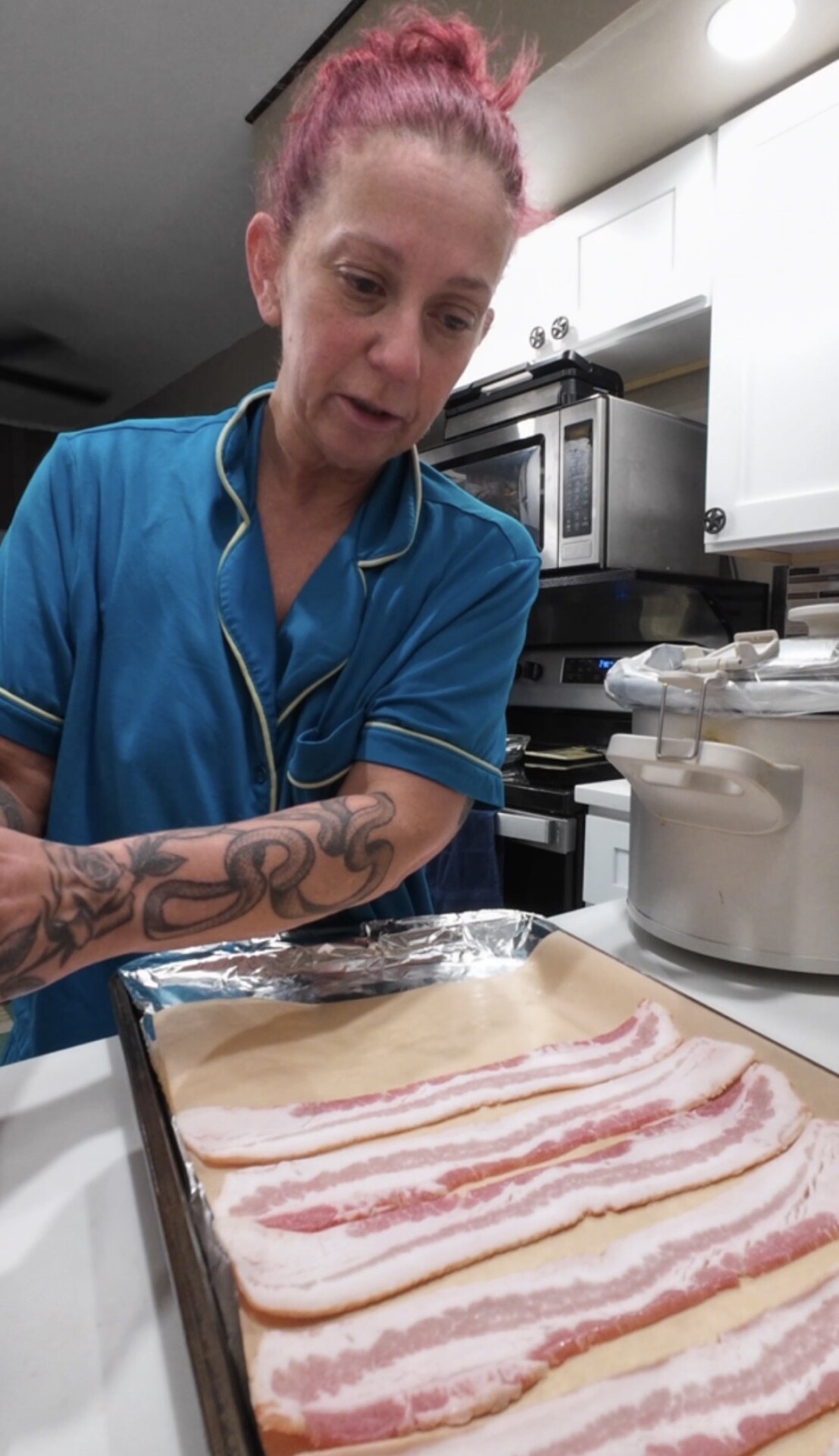 A person in a blue shirt with a tattoo on their arm arranging raw bacon strips on a baking sheet lined with parchment paper.