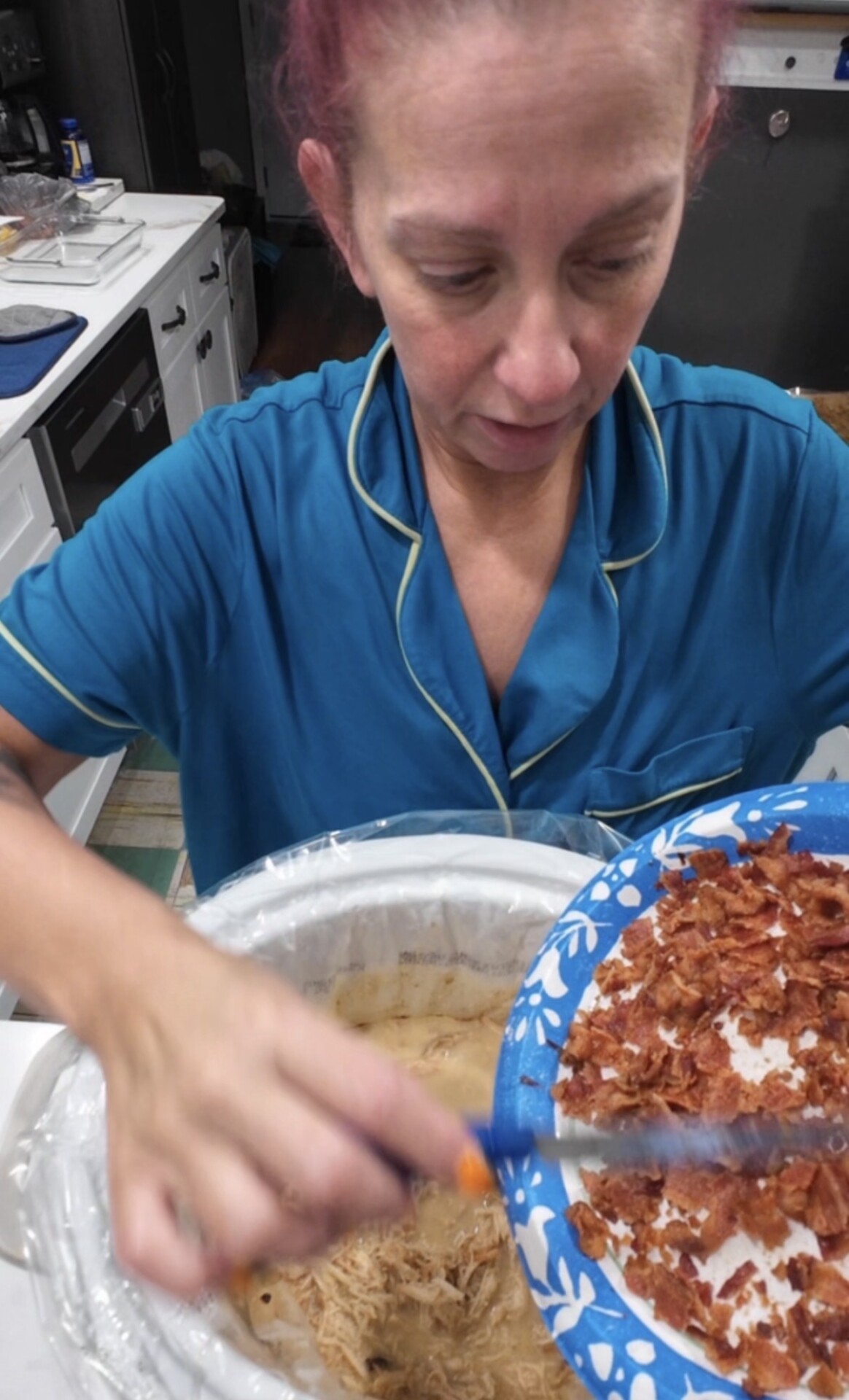 A person in a blue pajama top scrapes cooked, crumbled bacon from a patterned paper plate into a slow cooker containing shredded meat.