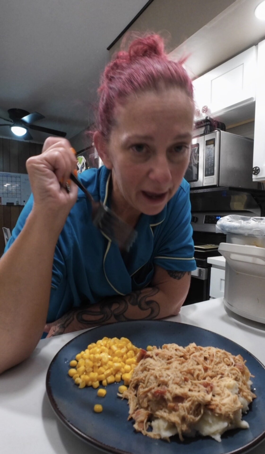 A person with pink hair holding a fork, sitting at a counter in front of a plate with corn and shredded meat over mashed potatoes.