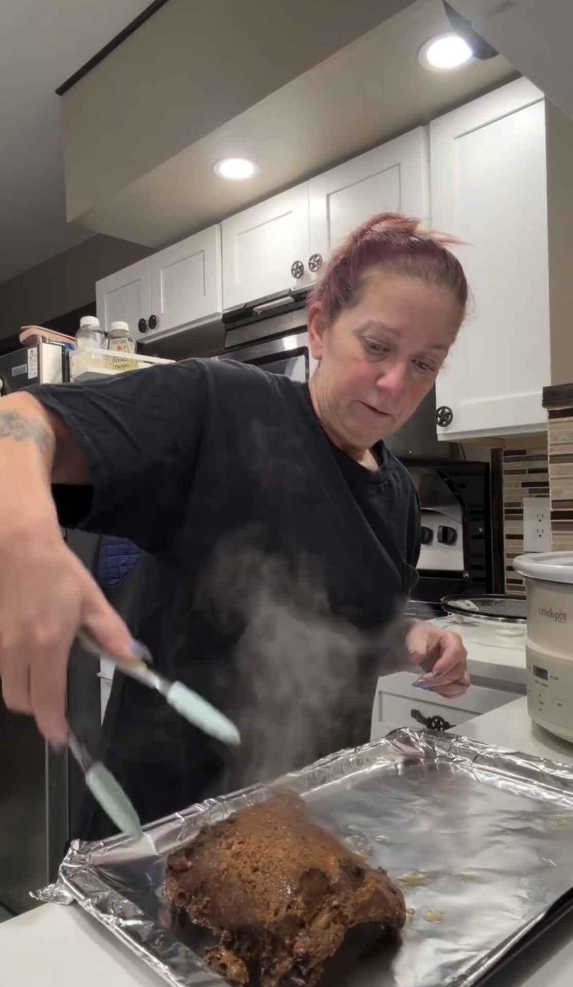 A woman uses kitchen tongs to handle a steaming piece of cooked meat on a foil-lined baking sheet.
