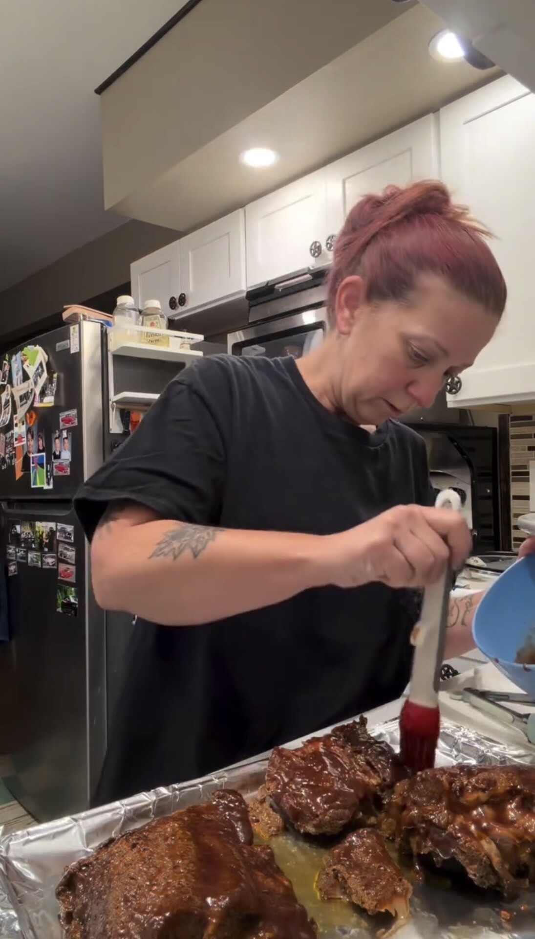 A person brushes barbecue sauce onto a tray of ribs in a kitchen.