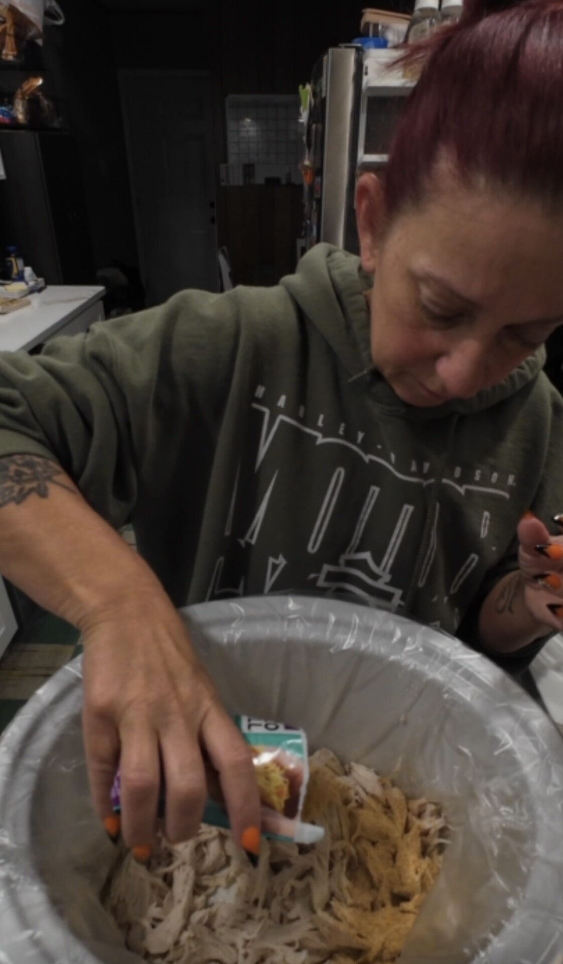 A person in a green hoodie pours a packet of seasoning into a slow cooker filled with shredded chicken.
