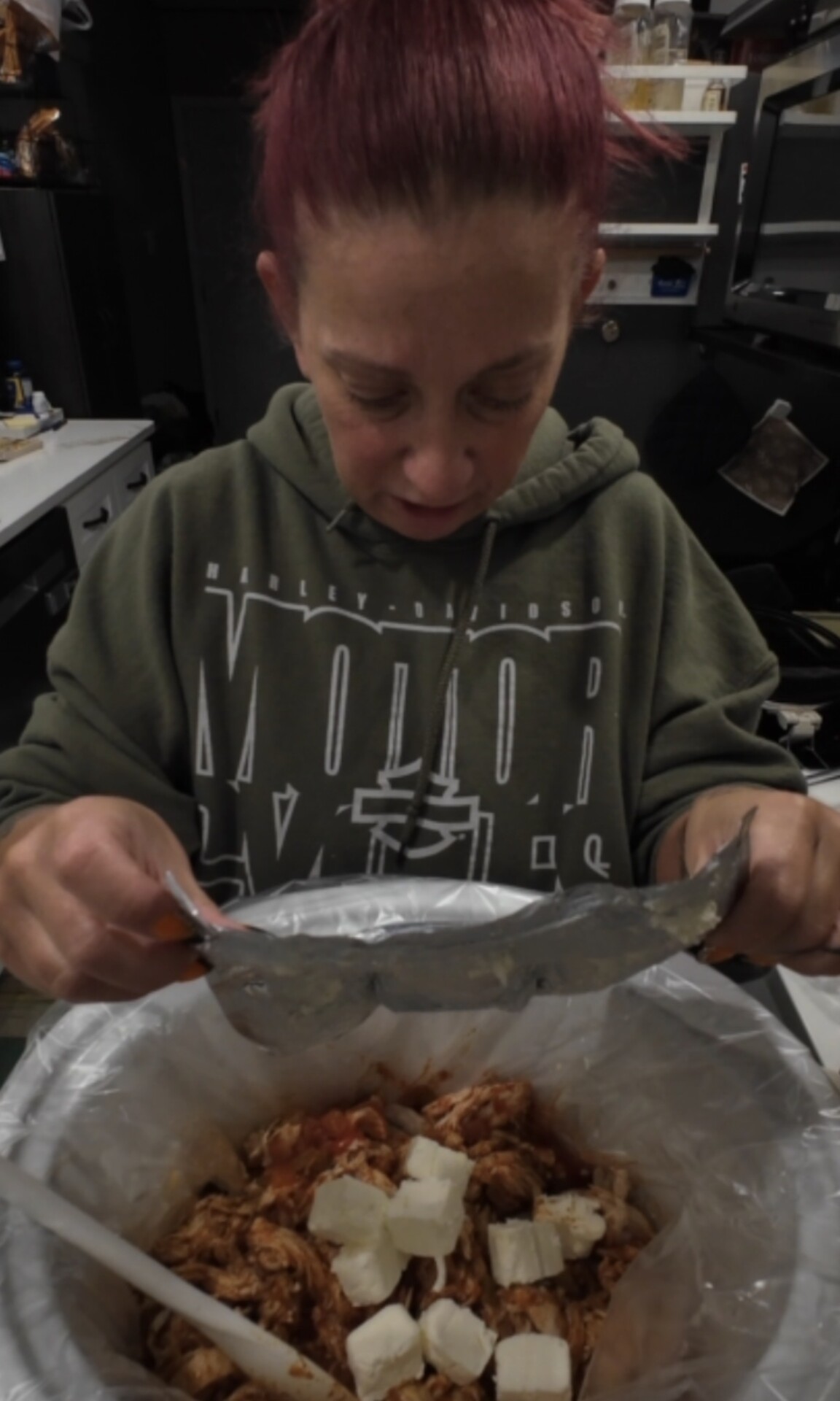 A person in an olive green hoodie adds a packet of ingredients to a slow cooker filled with shredded chicken and cubes of cream cheese.