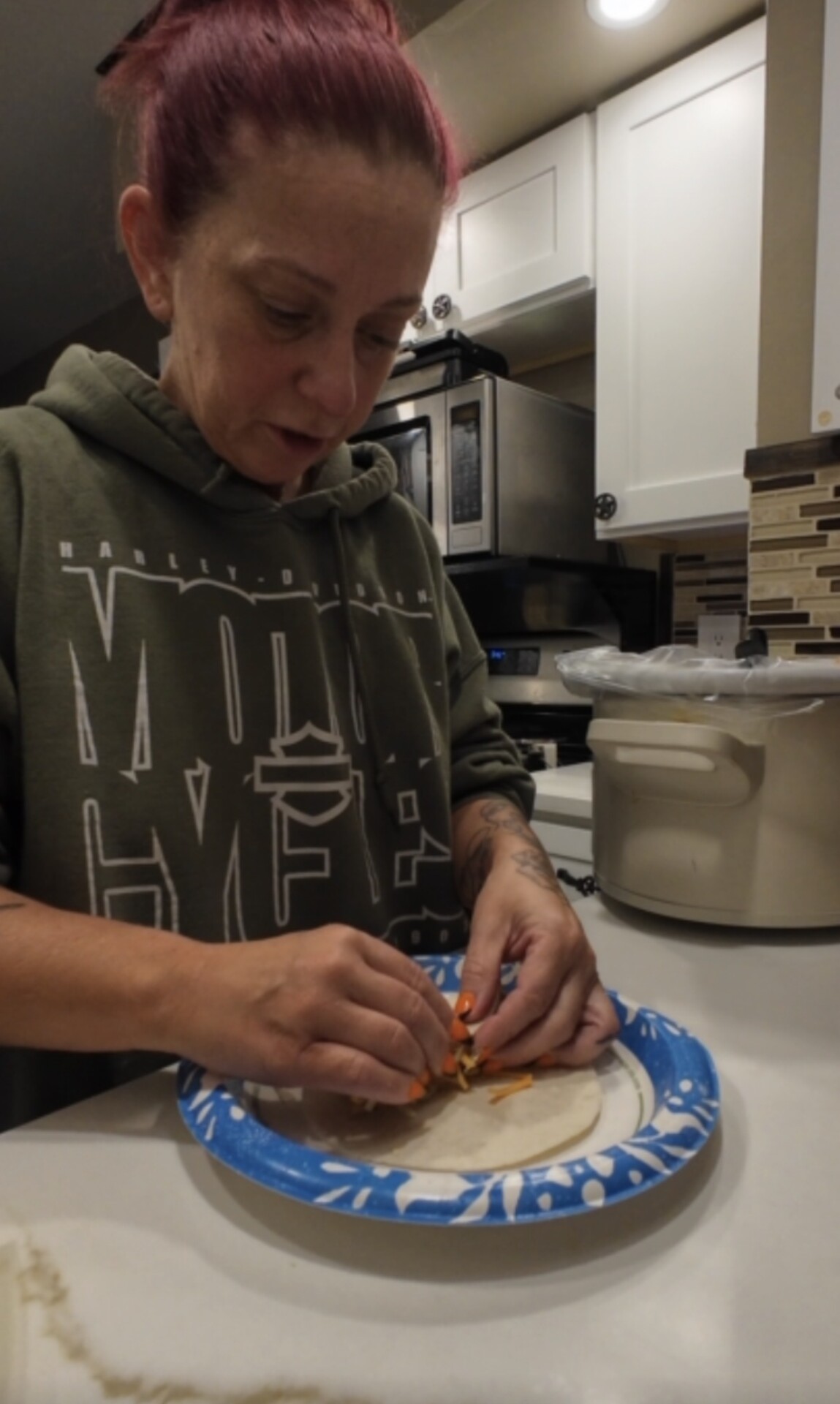 A person wearing an olive green hoodie places shredded cheese and vegetables onto a flour tortilla on a paper plate.