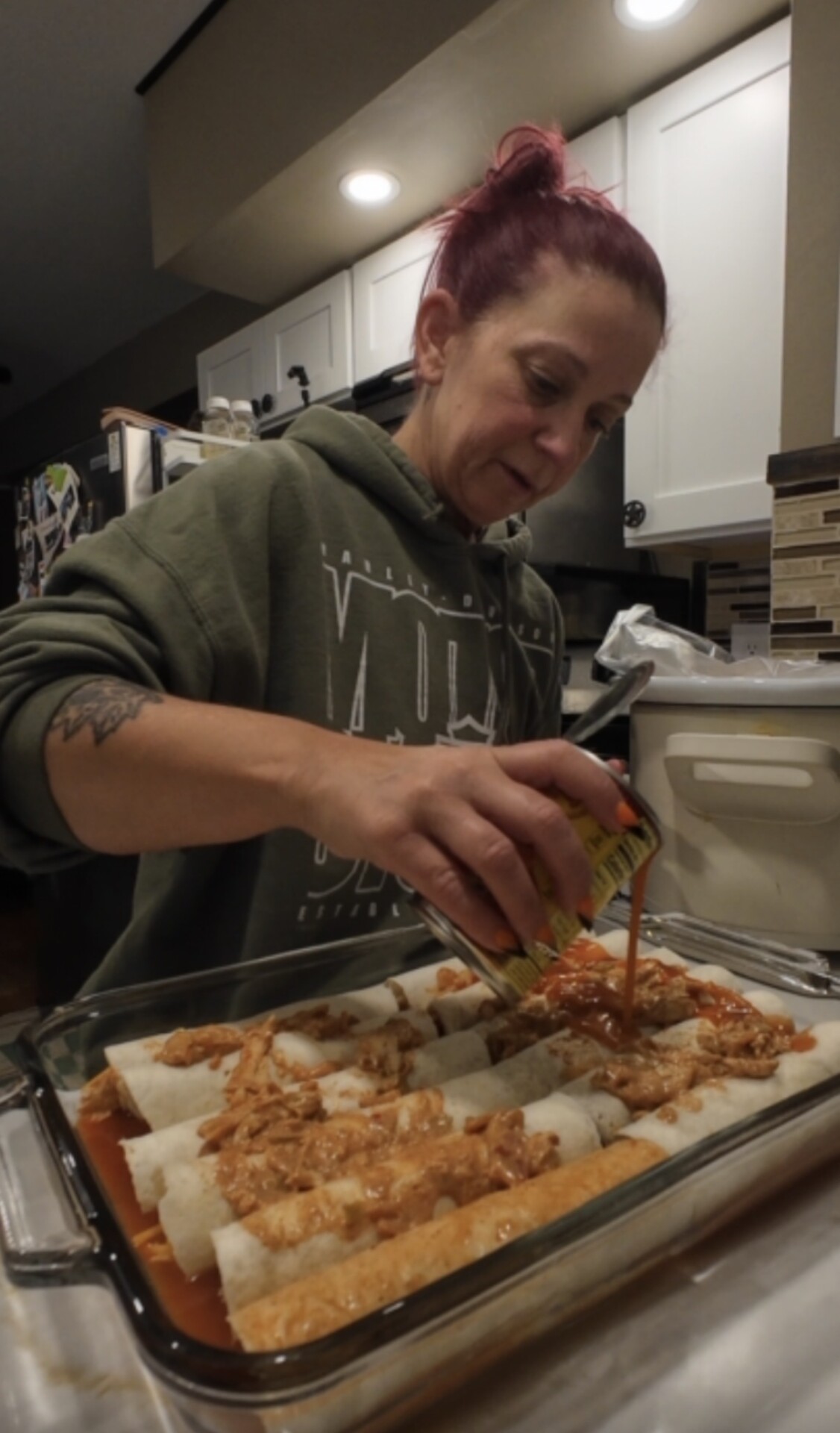 A woman in a green hoodie pours red sauce from a can over a row of chicken enchiladas in a glass baking dish.