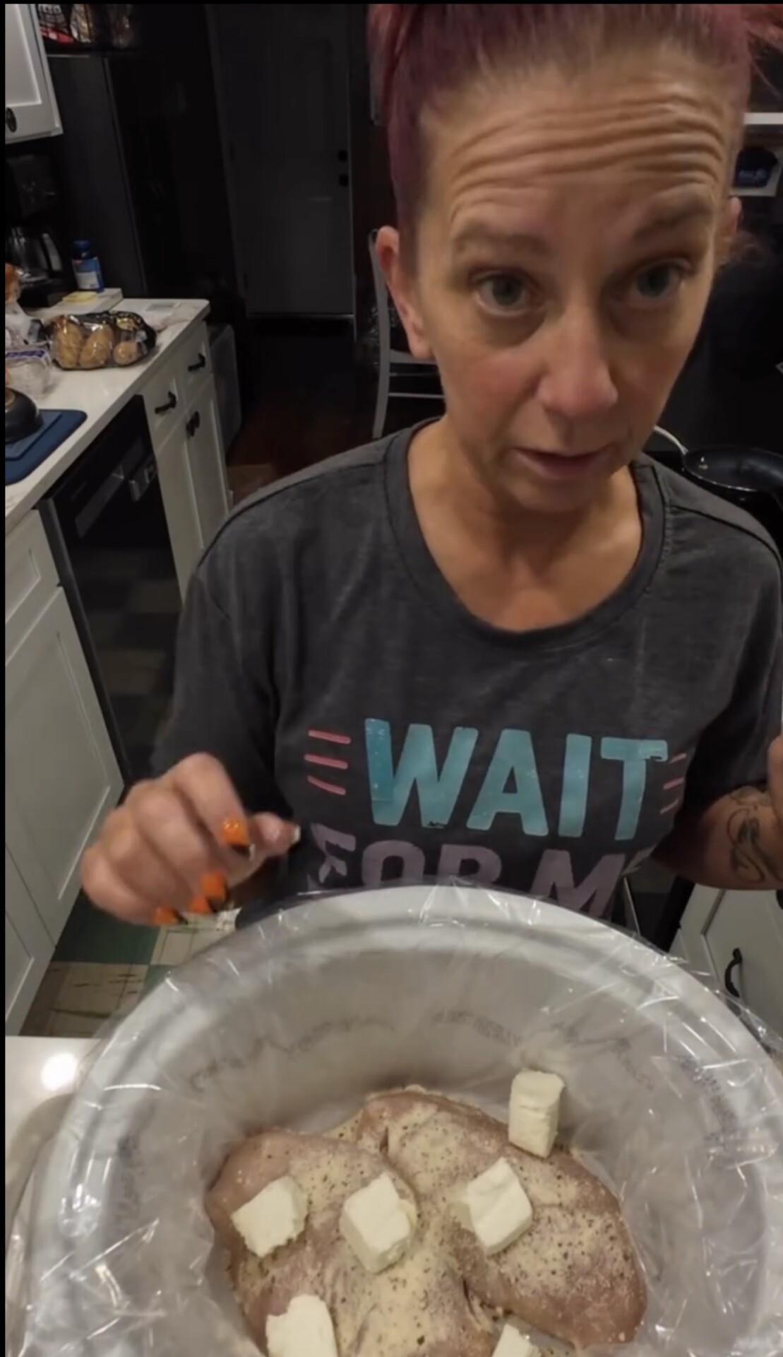 A person in a gray graphic t-shirt prepares chicken breasts in a slow cooker lined with a plastic liner, adding cubes of butter to the seasoned meat.