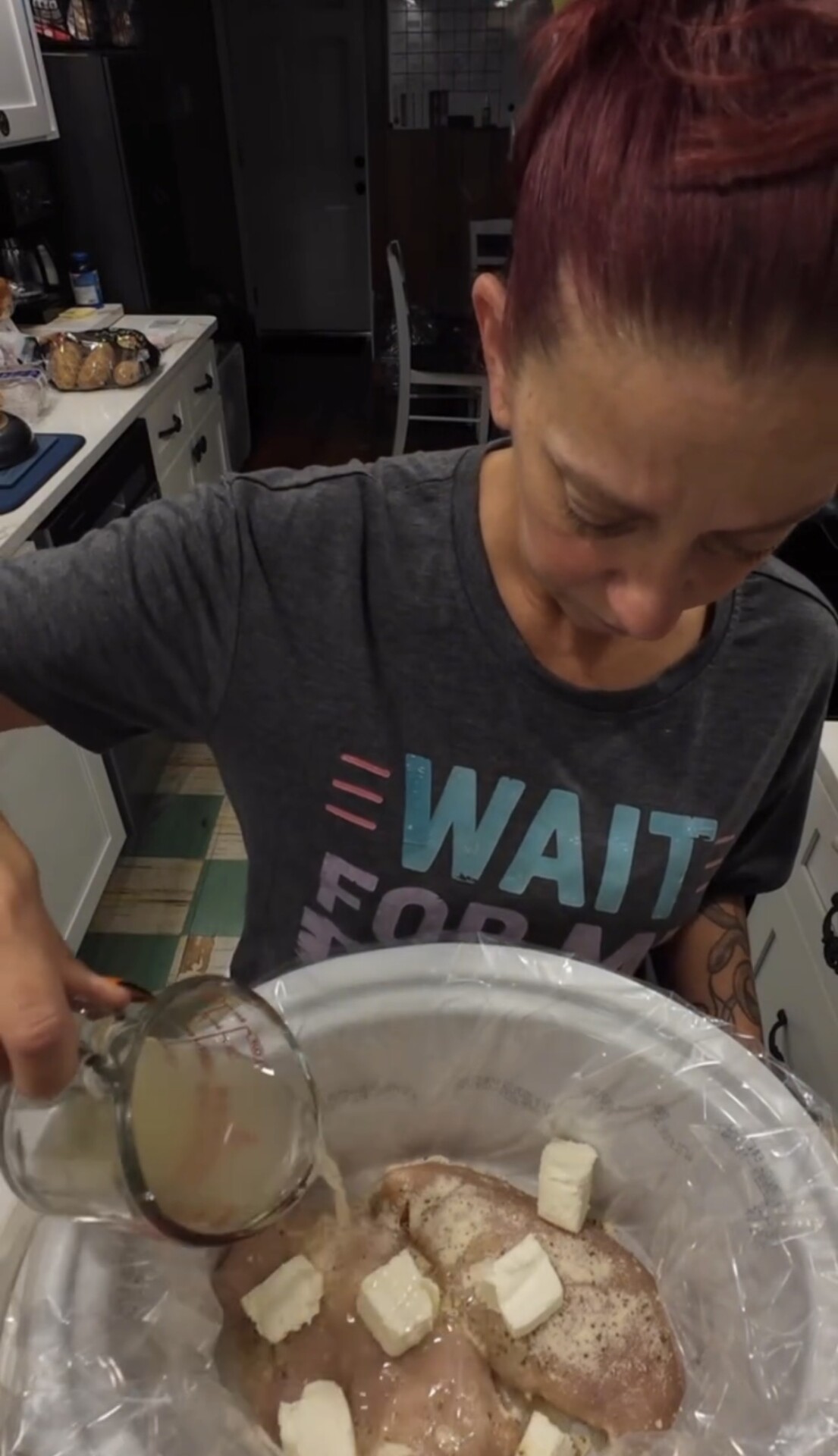 A person pours liquid from a measuring cup into a slow cooker containing seasoned chicken breasts and pats of butter.