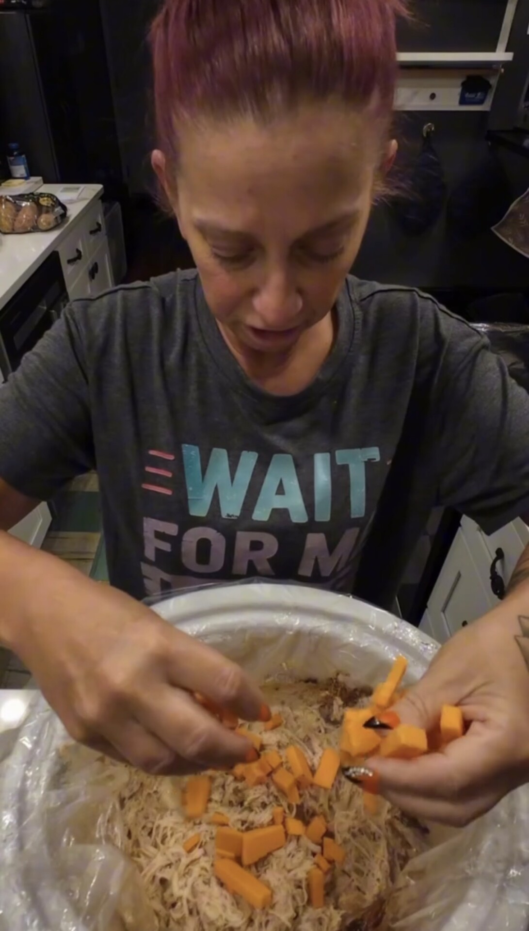 A person in a gray shirt adding chopped orange vegetables to a slow cooker filled with shredded meat.