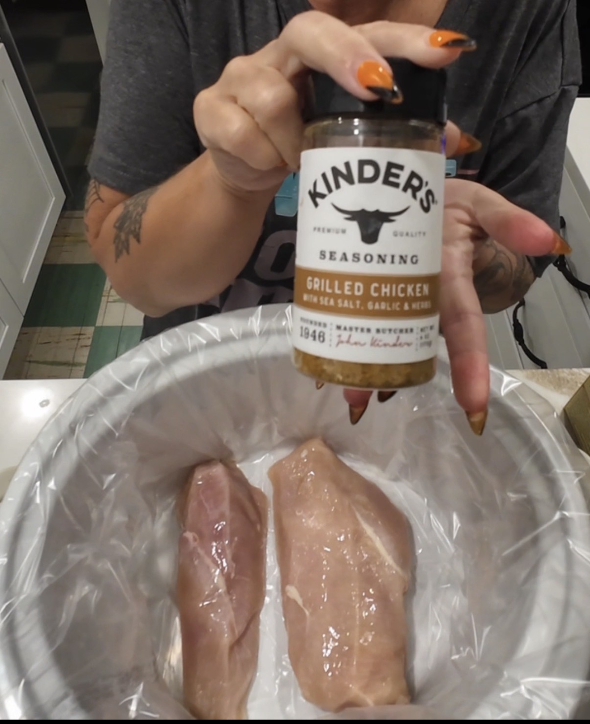 A person holds a bottle of Kinders Grilled Chicken seasoning over two raw chicken breasts in a slow cooker lined with a plastic bag.