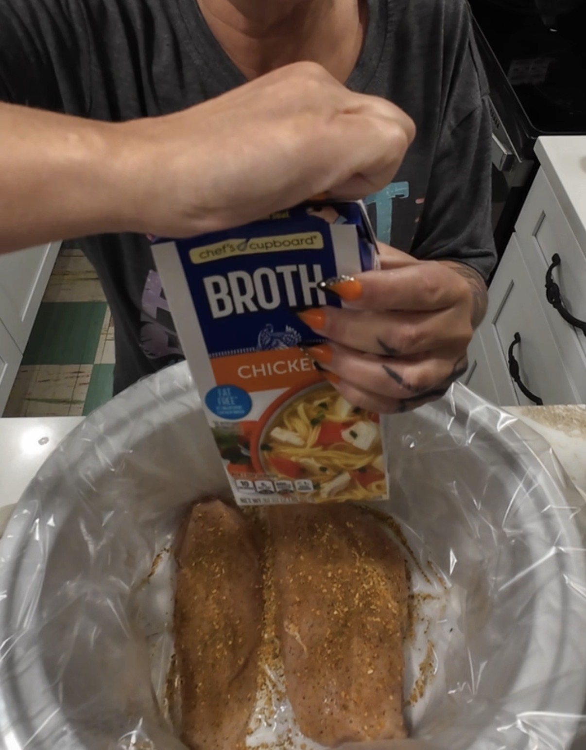 A person pouring a carton of chicken broth into a slow cooker over seasoned chicken breasts.