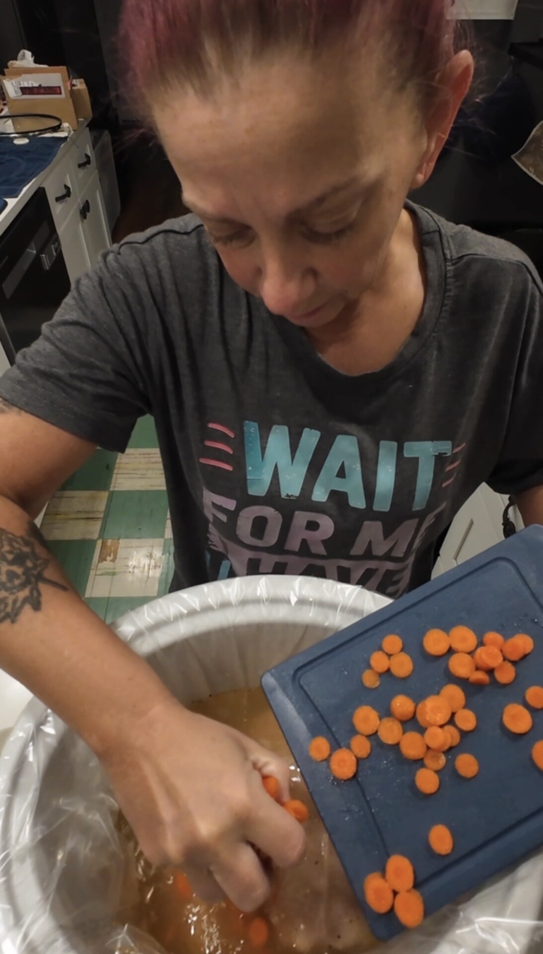 A person wearing a grey shirt empties sliced carrots from a blue cutting board into a slow cooker.