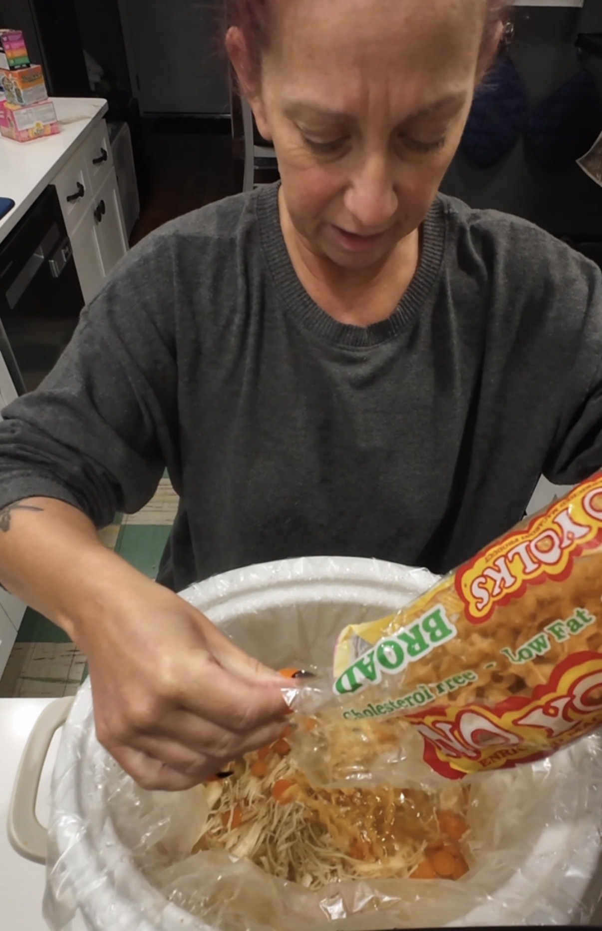 A person pouring a bag of Broad Yolks egg noodles into a slow cooker containing shredded chicken and carrots.