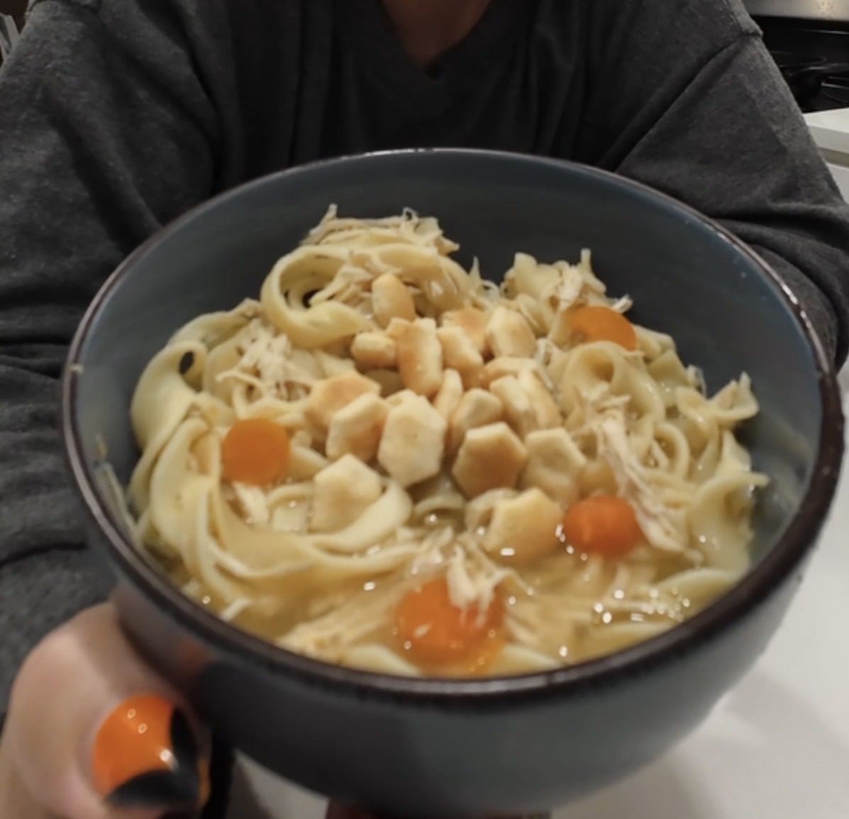 A dark bowl of chicken noodle soup topped with oyster crackers, held by a person in a gray shirt.
