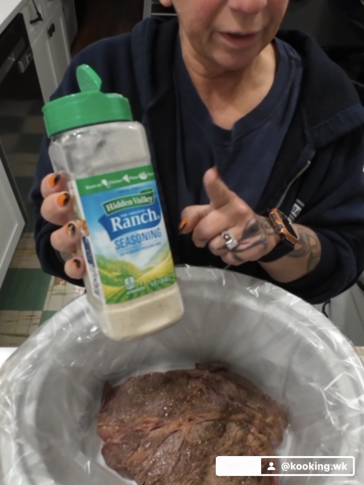 A person holding a container of Hidden Valley Ranch seasoning over a slow cooker containing a large piece of meat.