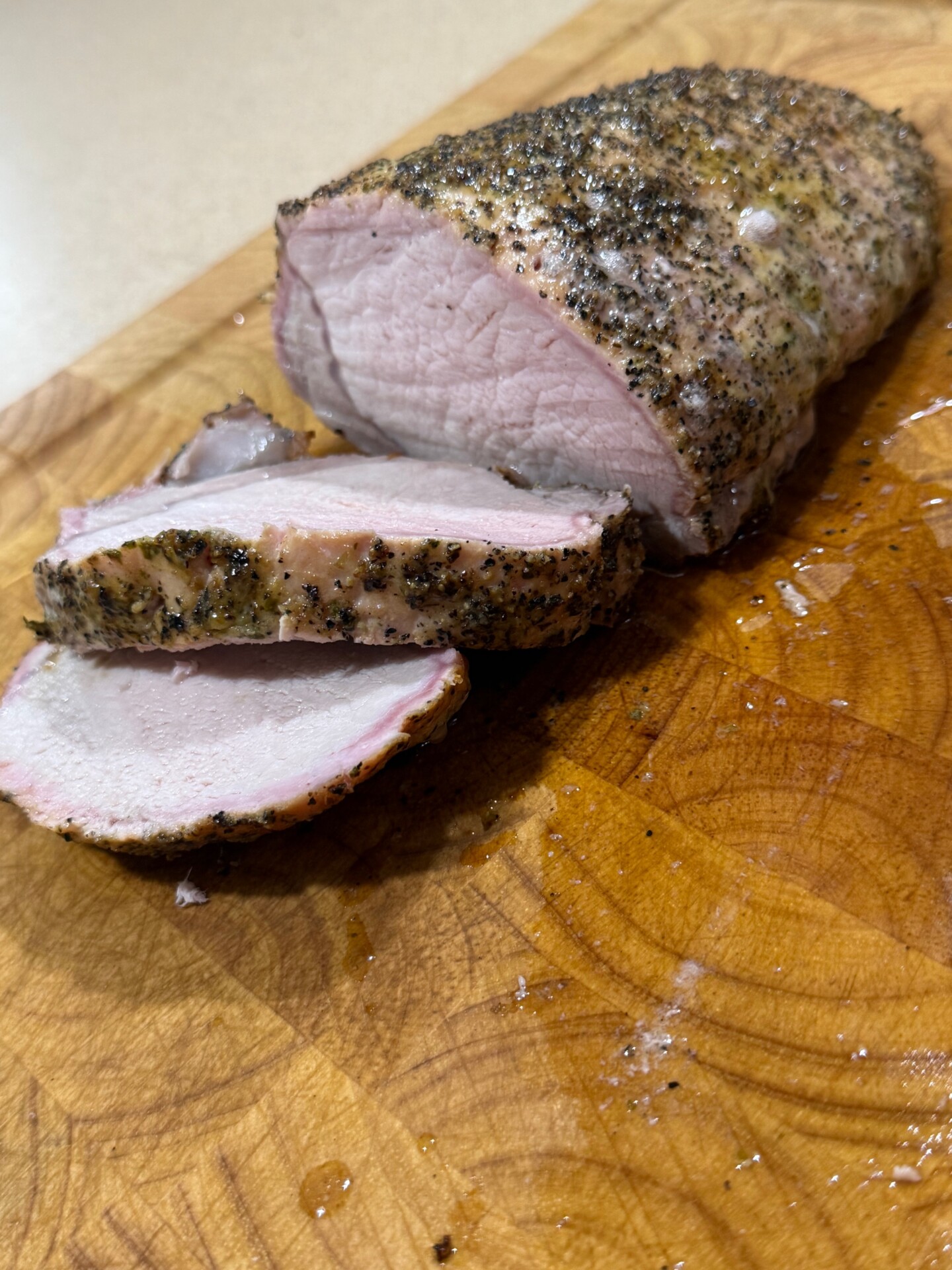 Sliced, cooked pork tenderloin with a peppery crust sits on a wooden cutting board, showing juicy and tender meat inside.