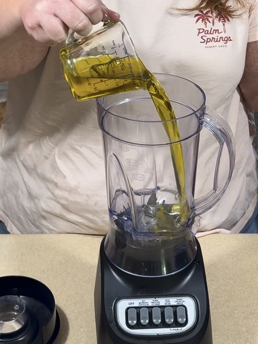 A person pours yellow liquid, possibly oil, from a measuring cup into a clear blender. The person is wearing a light pink Palm Springs t-shirt. The blender sits on a countertop.