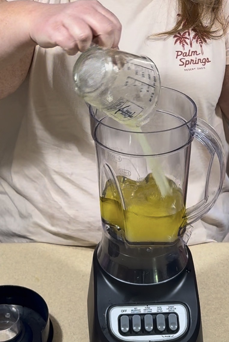 A person pours liquid from a measuring cup into a blender containing a yellow substance. The blender sits on a countertop, and the person is wearing a shirt with a Palm Springs logo.