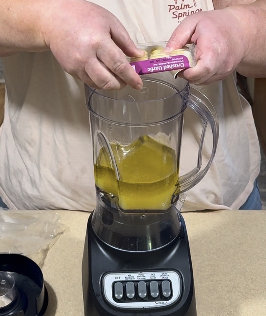 A person adds crushed garlic to a blender containing yellow liquid, possibly oil. The blender is on a countertop, and the person is wearing a light pink shirt.