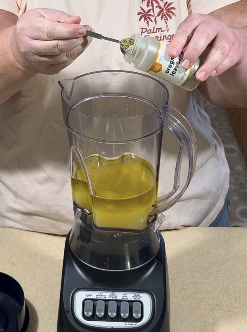 A person adds dried oregano from a spice bottle into a blender filled with a yellow liquid, possibly oil. The blender is on a countertop, and the person is wearing a palm tree patterned shirt.