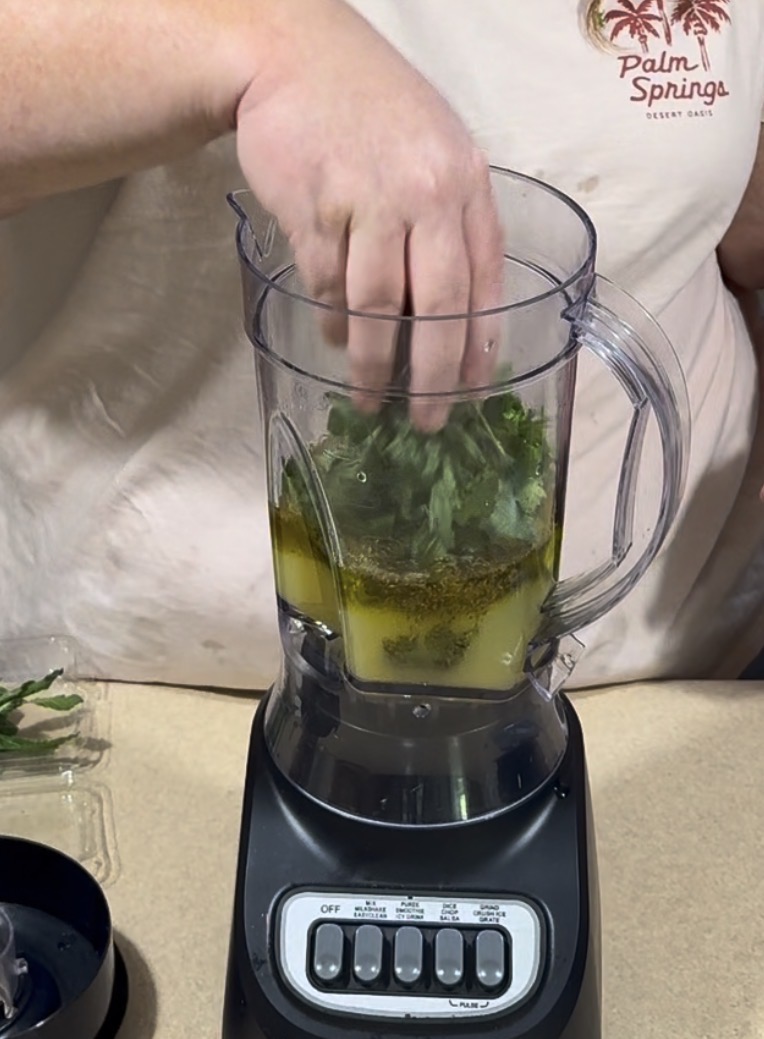 A person places fresh herbs into a blender filled with oil and seasonings. The blender is on a countertop, and the person is wearing a Palm Springs t-shirt.