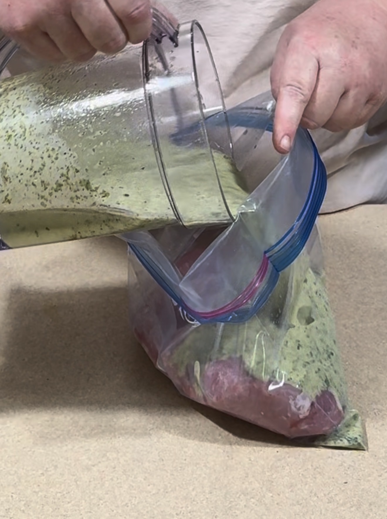 A person pours green marinade from a blender into a zip-top bag containing a raw piece of meat, preparing it for marinating on a beige countertop.