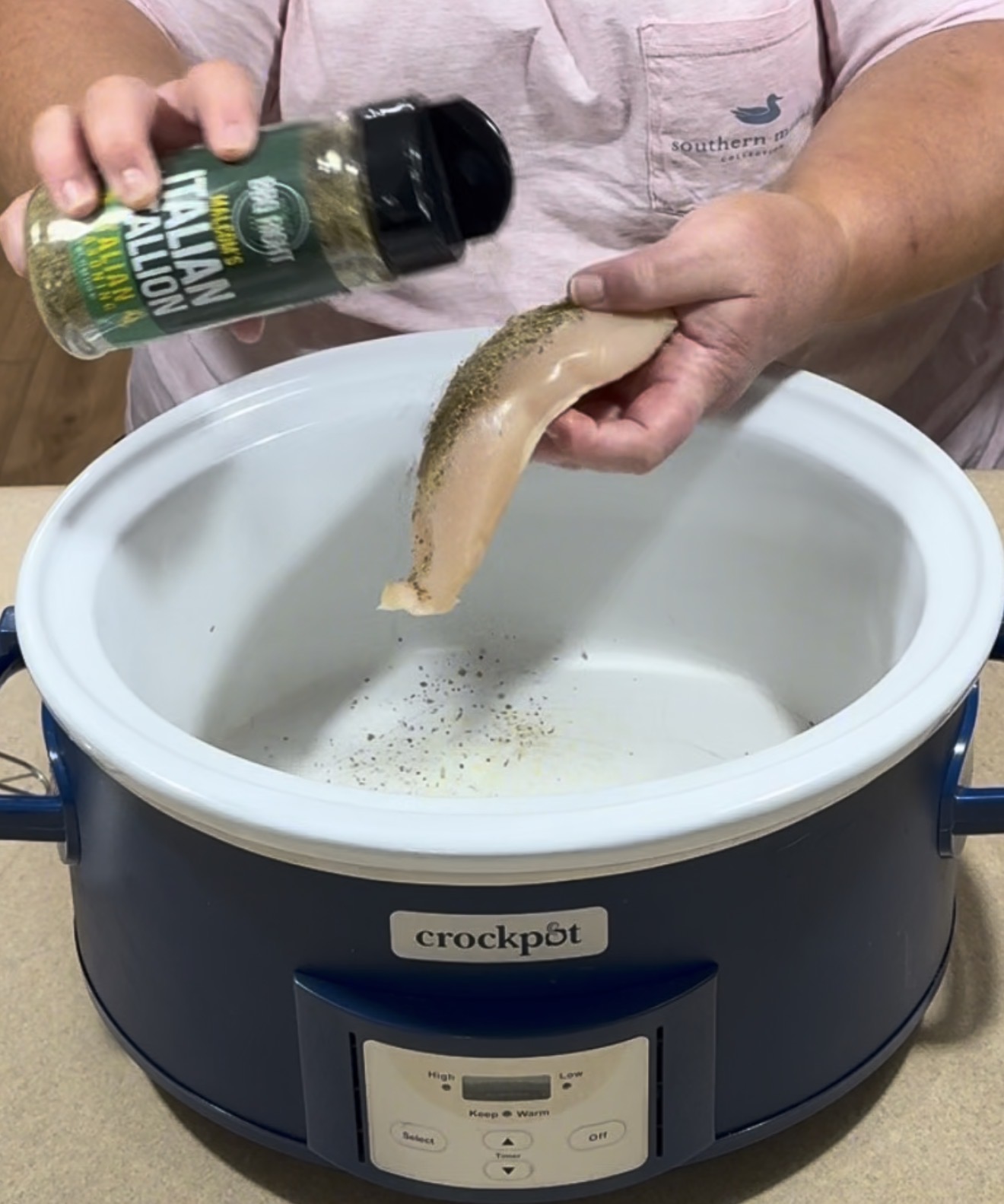 A person sprinkles Italian seasoning onto a raw chicken breast over a blue slow cooker labeled crockpot.