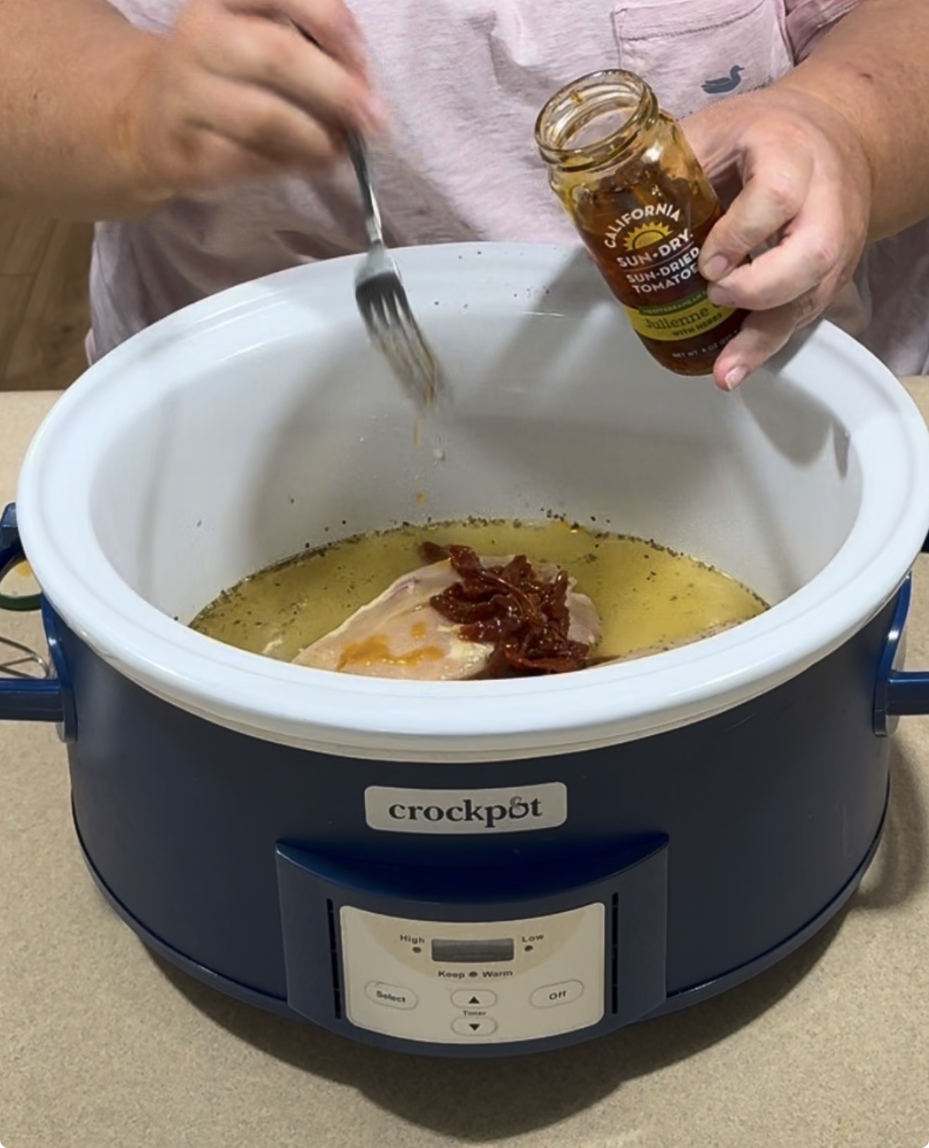 A person adds sun dried tomatoes from a jar into a crockpot containing chicken and broth, using a fork. The crockpot is blue and white, placed on a countertop.