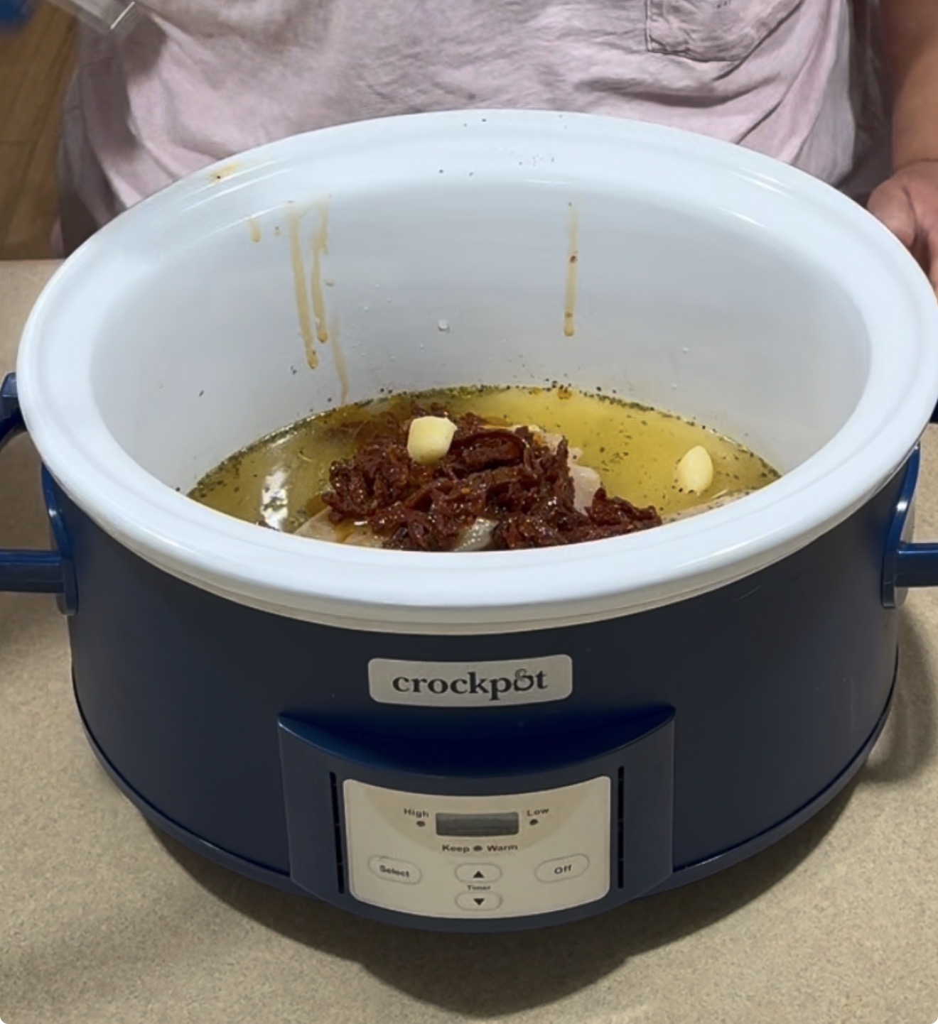 A person stands behind a blue Crock-Pot filled with oil, sun-dried tomatoes, garlic cloves, and spices, preparing ingredients for cooking.