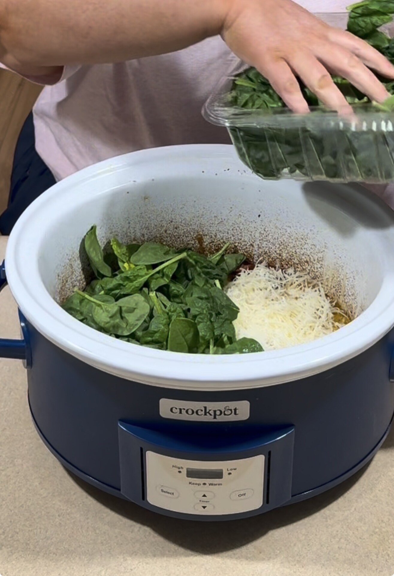 A person adds fresh spinach from a container into a blue Crock-Pot filled with shredded cheese and other ingredients, preparing a meal.