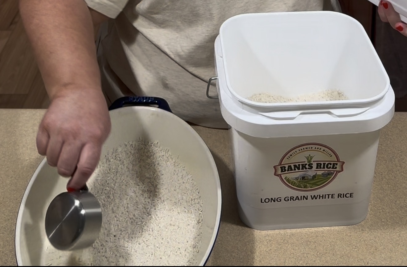 A person uses a measuring cup to pour Banks Rice long grain white rice from a plastic container into a baking dish.